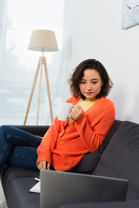 young freelancer holding cup and looking at laptop on couch