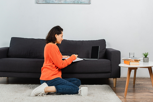 woman sitting on carpet and writing in notebook near laptop with blank screen on couch
