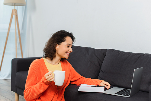 happy young woman holding cup of coffee while looking at laptop