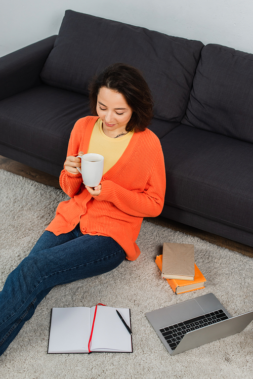 high angle view of tattooed young woman sitting on carpet and holding cup of coffee near laptop and books