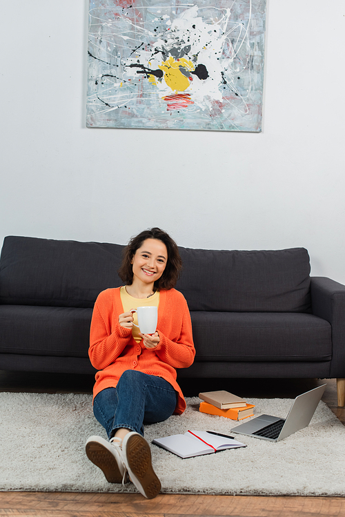 happy young woman sitting on carpet and holding cup of coffee near laptop and books