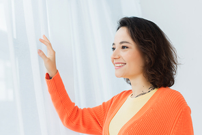 tattooed and pleased young woman smiling near white curtain