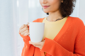 cropped view of tattooed young woman holding cup near white curtain