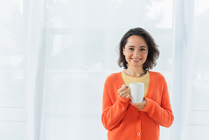 positive young woman smiling and holding cup near white curtain