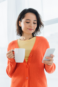 tattooed young woman holding cup and smartphone near white curtain