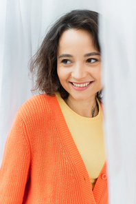 portrait of positive young woman with tattoo smiling near white curtain