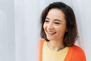 portrait of joyful and tattooed young woman smiling near white curtain