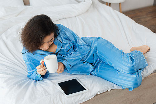 high angle view of woman in blue pajamas holding cup of coffee and using digital tablet in bedroom