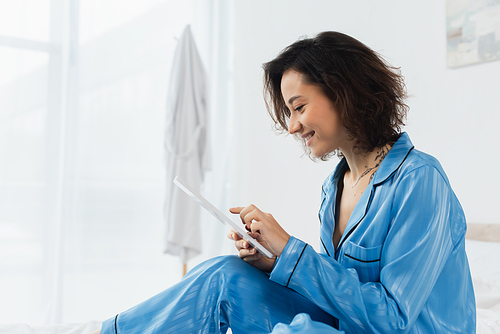 happy young woman in blue pajamas using digital tablet at home
