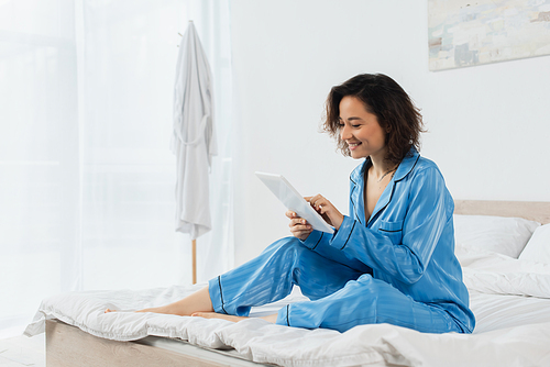 cheerful young woman in blue pajamas using digital tablet in bedroom