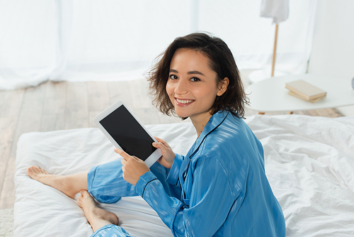 high angle view of happy young woman in blue pajamas using digital tablet in bedroom