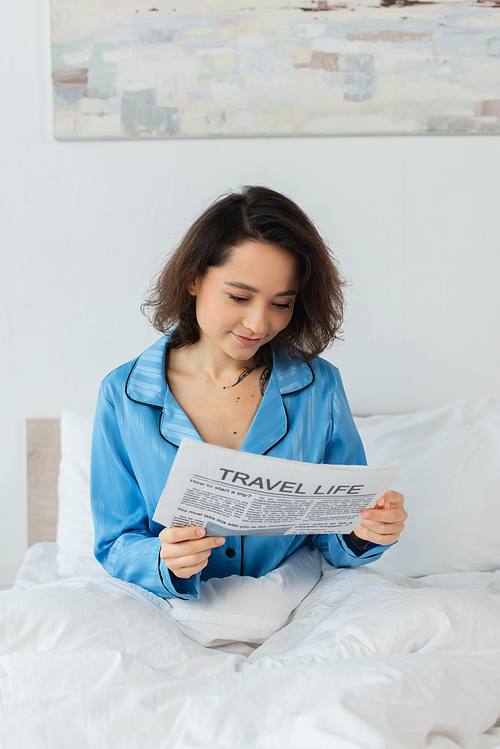 brunette woman in blue pajamas reading travel life newspaper in bed