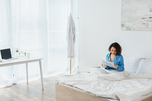 brunette woman in blue pajamas reading travel life newspaper in bed near laptop on desk