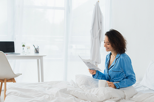 happy woman in blue pajamas reading travel life newspaper and holding cup in bed