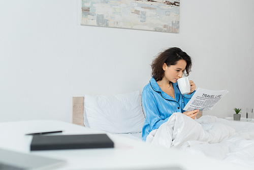 brunette woman in blue pajamas reading travel life newspaper and drinking . in bed
