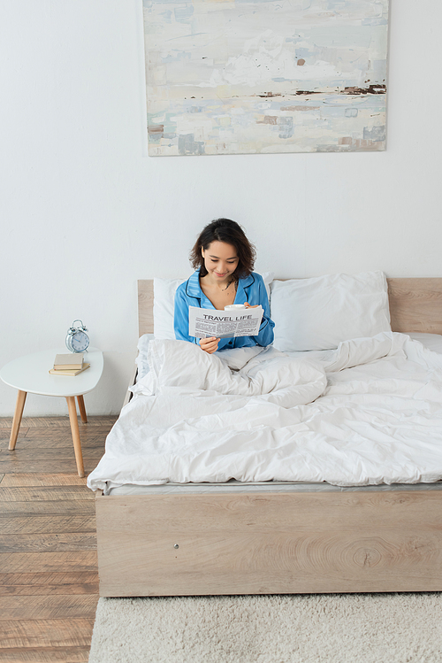 cheerful young woman in pajamas reading travel life newspaper and holding cup in bed