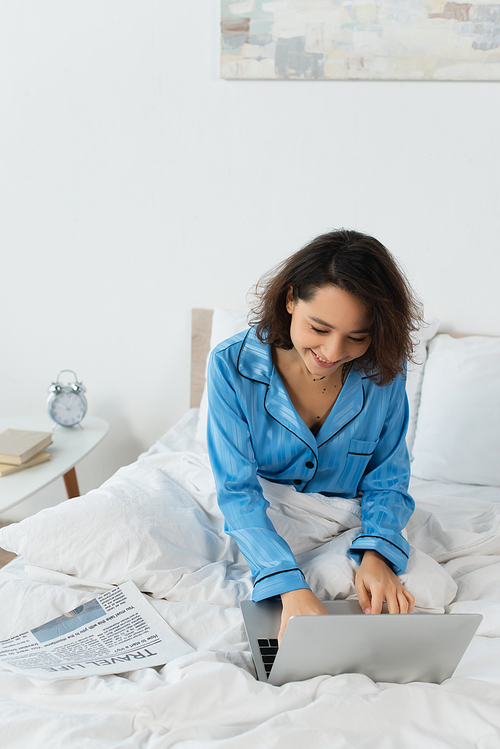 smiling young woman in pajamas using laptop near newspaper on bed