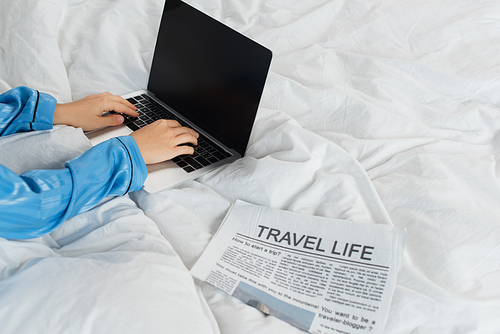 cropped view of young woman in pajamas using laptop near newspaper on bed