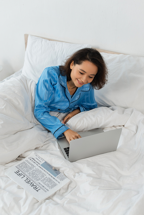 smiling young freelancer in pajamas using laptop near newspaper on bed