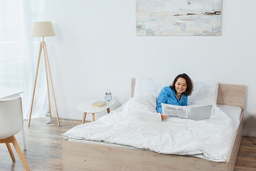 cheerful young woman in pajamas reading newspaper near laptop on bed