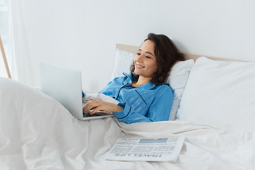 cheerful young freelancer in blue pajamas using laptop near newspaper on bed