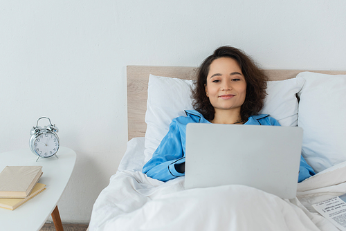 positive young freelancer in blue pajamas using laptop in bed