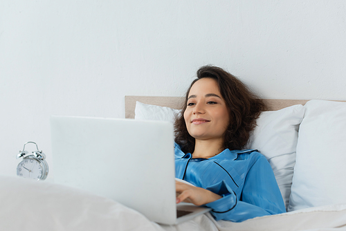 cheerful young woman in pajamas using laptop near alarm clock in bedroom