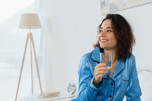 happy young woman in blue pajamas holding glass of water in bedroom