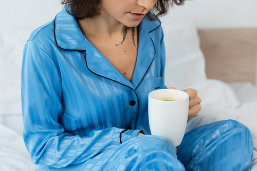 cropped view of tattooed young woman in blue pajamas holding cup of coffee in bedroom