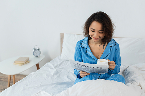 cheerful young woman in pajamas reading travel life newspaper and holding cup in bed