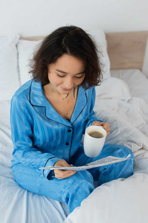 high angle view of young woman in blue pajamas holding cup of coffee while reading newspaper in bedroom