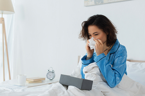 sick woman sneezing in napkin near tissue box on bed