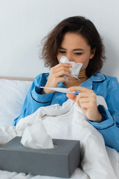 ill young woman sneezing in tissue and looking at electronic thermometer