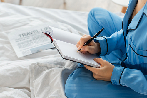 cropped view of young woman writing in notebook near newspaper on bed