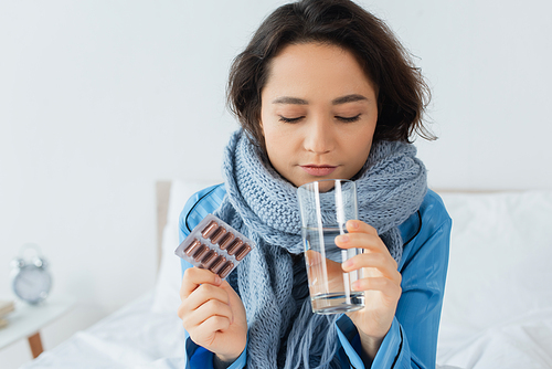 sick young woman in knitted scarf holding glass of water and blister pack with pills