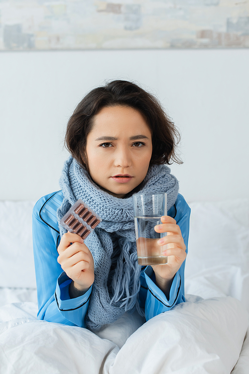 sick woman in knitted scarf holding glass of water and blister pack with capsules