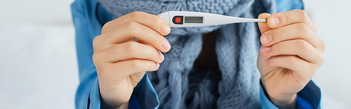 cropped view of sick woman in knitted scarf holding electronic thermometer, banner
