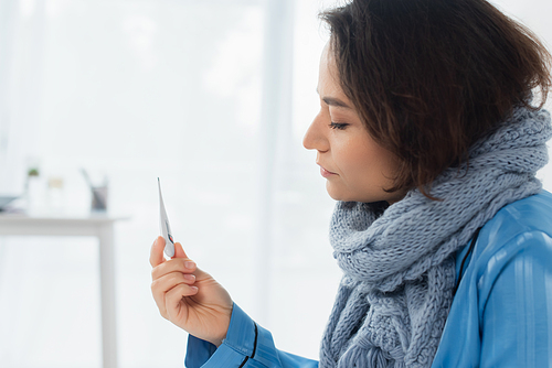 sick young woman in blue knitted scarf holding electronic thermometer