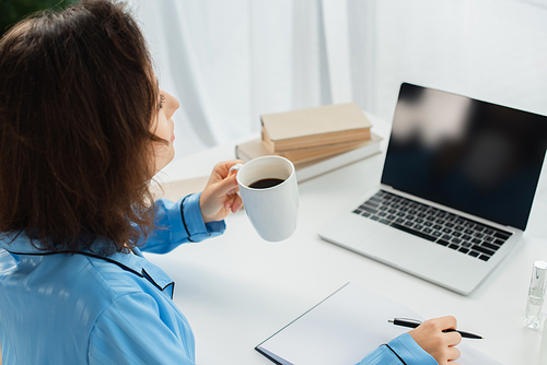 brunette woman holding cup of coffee near laptop with blank screen on desk