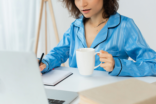 cropped view of young woman holding cup of coffee near notebook and laptop