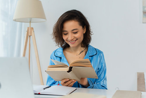 happy young woman reading book near laptop on desk