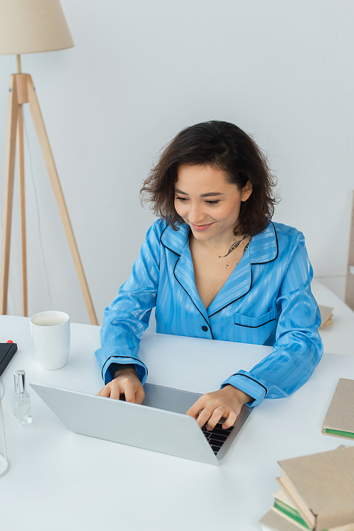 cheerful young freelancer using laptop near cup and books