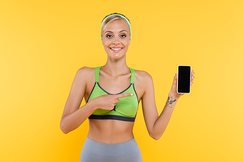 joyful woman in sportswear pointing at mobile phone with blank screen isolated on yellow
