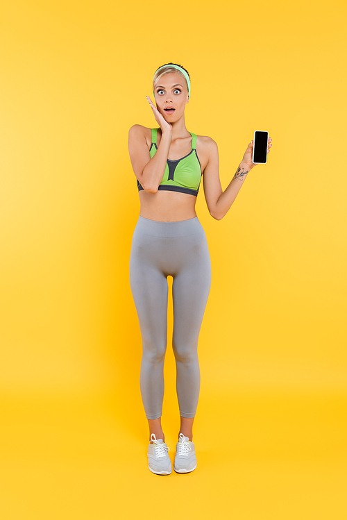 full length view of amazed woman in sportswear touching face while showing smartphone with blank screen on yellow