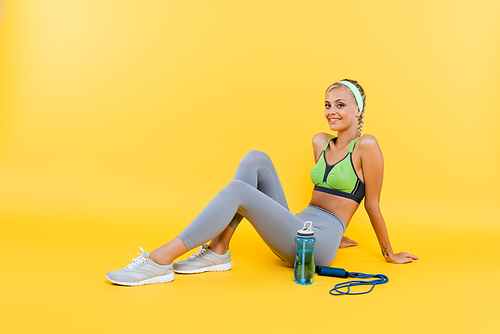 cheerful woman in sportswear  while sitting near sports bottle and skipping rope on yellow