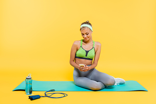 pleased woman in sportswear using smartphone while sitting on fitness mat near sports bottle and jump rope on yellow