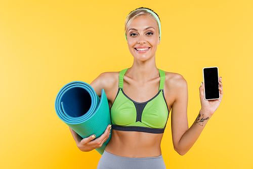 happy woman in green sports bra holding fitness mat while showing mobile phone with blank screen isolated on yellow