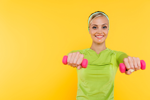 slim woman in green long sleeve shirt smiling while working out with dumbbells isolated on yellow