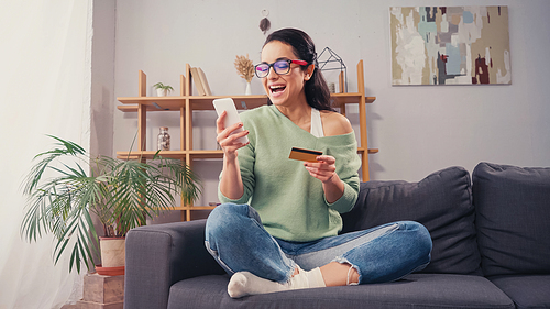 Excited woman in eyeglasses using smartphone and credit card