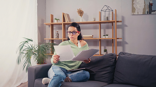 Brunette student in eyeglasses looking at documents near paper folder at home